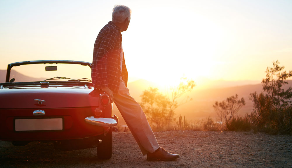 Grauhaariger Mann lehnt an einem roten Cabriolet und blickt in Richtung Sonnenuntergang